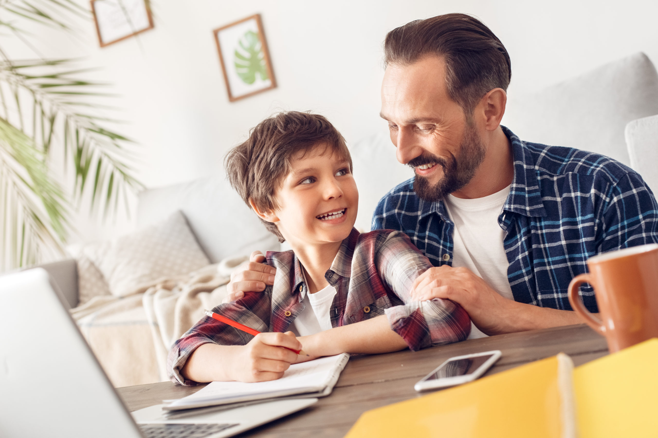 Father and little son together at home sitting at table caring dad helping son doing assignment smiling cheerful